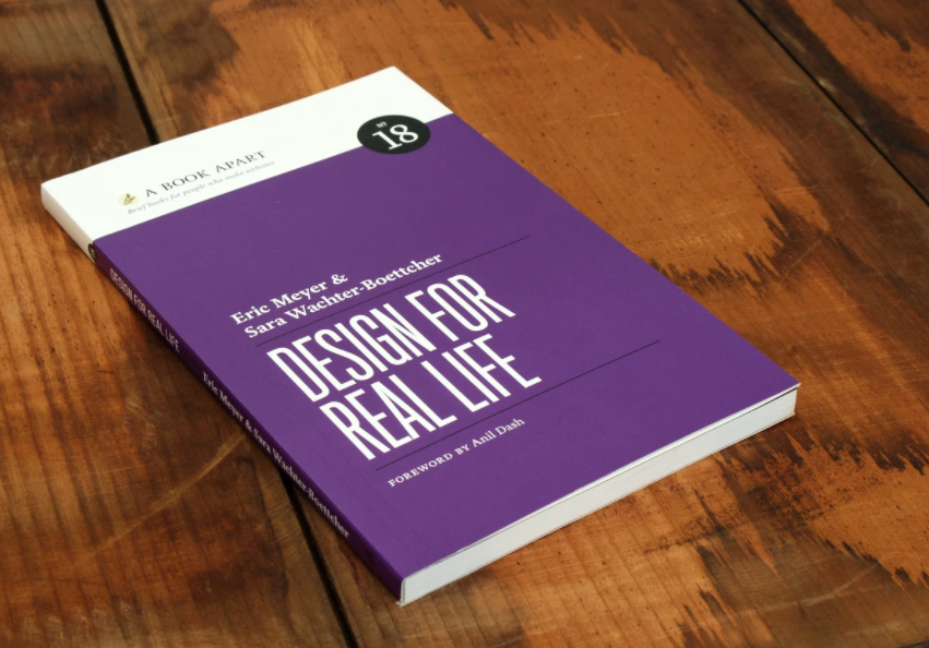 The cover of Design For Real Life. a simple purple cover, lying on a wooden surface