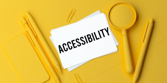 Pens, paperclips, a mobile phone, and a magnifying glass all spray painted yellow on a yellow surface, with a white paper that says ACCESSIBILITY in the centre
